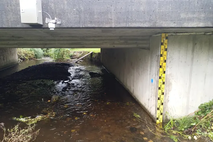 Pegelmessstelle an einem Bach unter einer Betonbrücke mit gelber Pegellatte